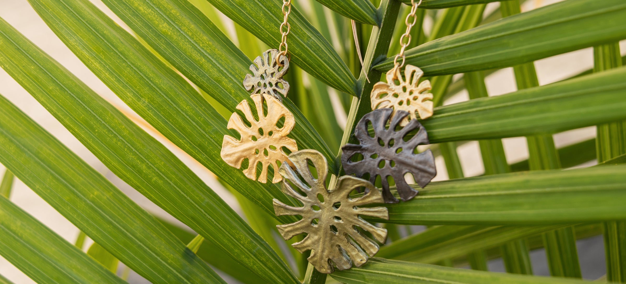 A necklace with gold and dark metal monstera leaf pendants hangs on green palm fronds.