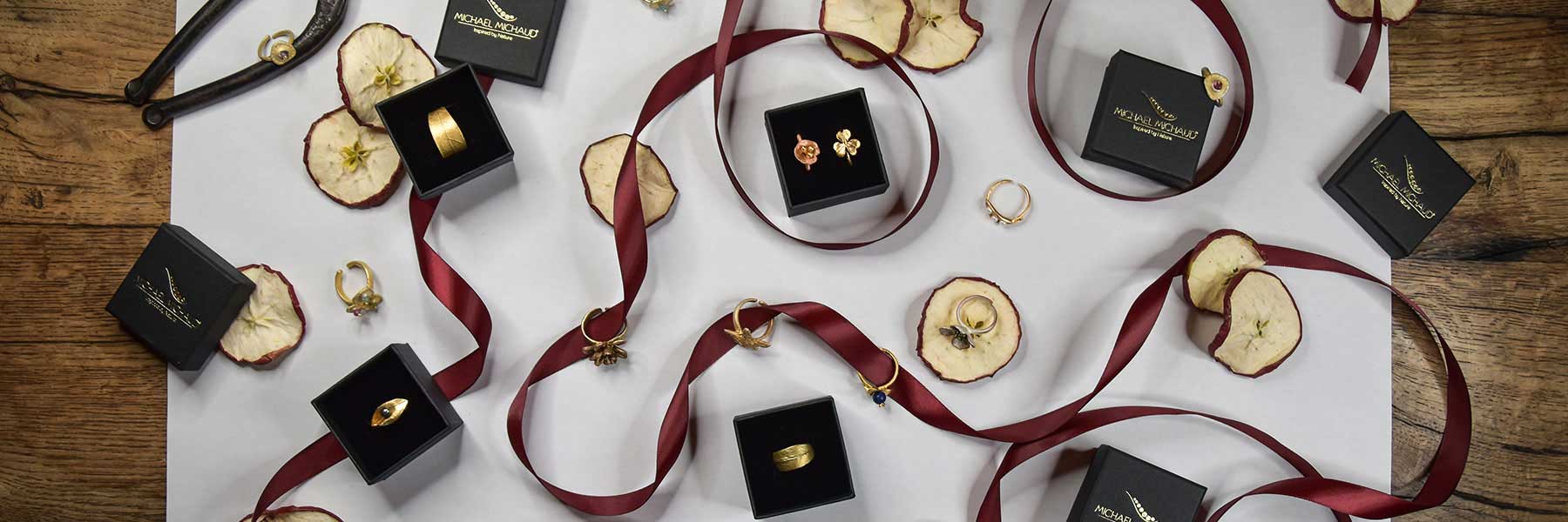 Gold jewelry pieces in black boxes, dried apple slices, and maroon ribbons arranged on a white and wooden surface.