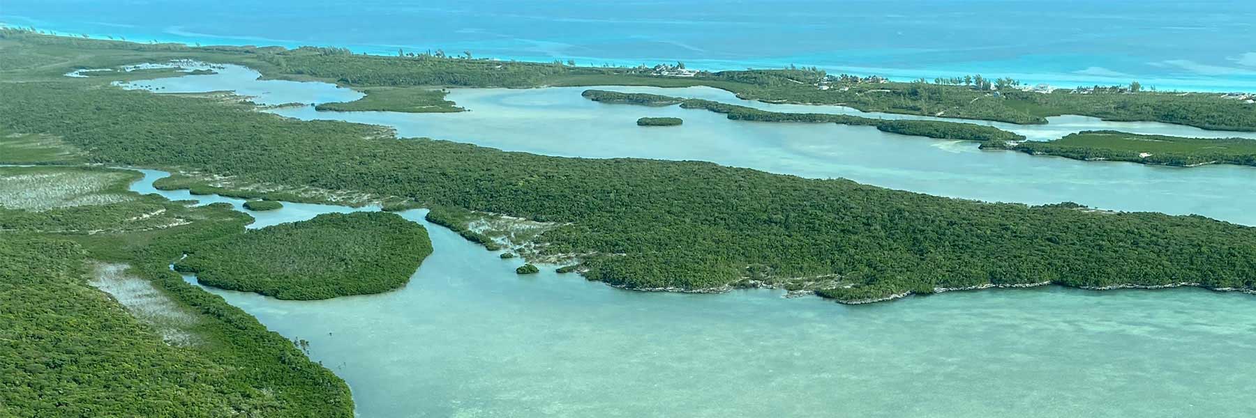 Aerial view of green islands and sandbars surrounded by shallow blue-green water, with a distant coastline and turquoise ocean in the background.