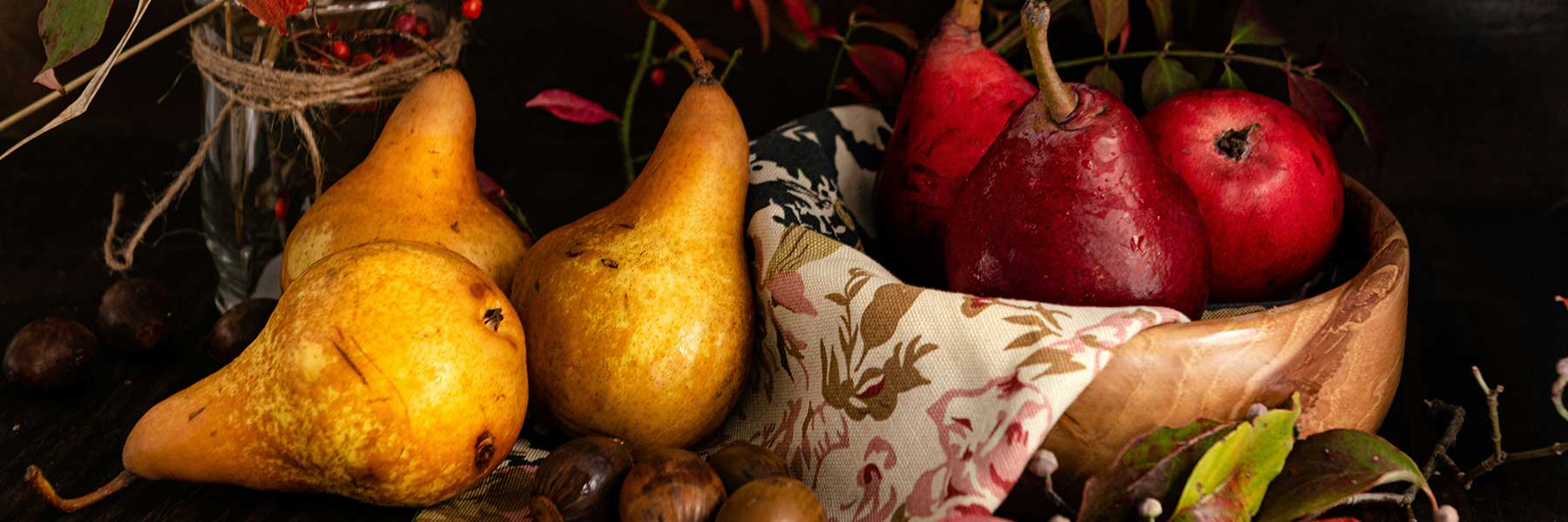 A wooden bowl holds red pears and an apple, while yellow pears and chestnuts are arranged beside it on a table with autumn leaves and a patterned cloth.