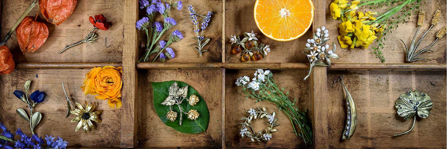 Wooden tray divided into sections, each containing various flowers, a half orange, green leaf, and an assortment of jewelry inspired by botanical shapes.
