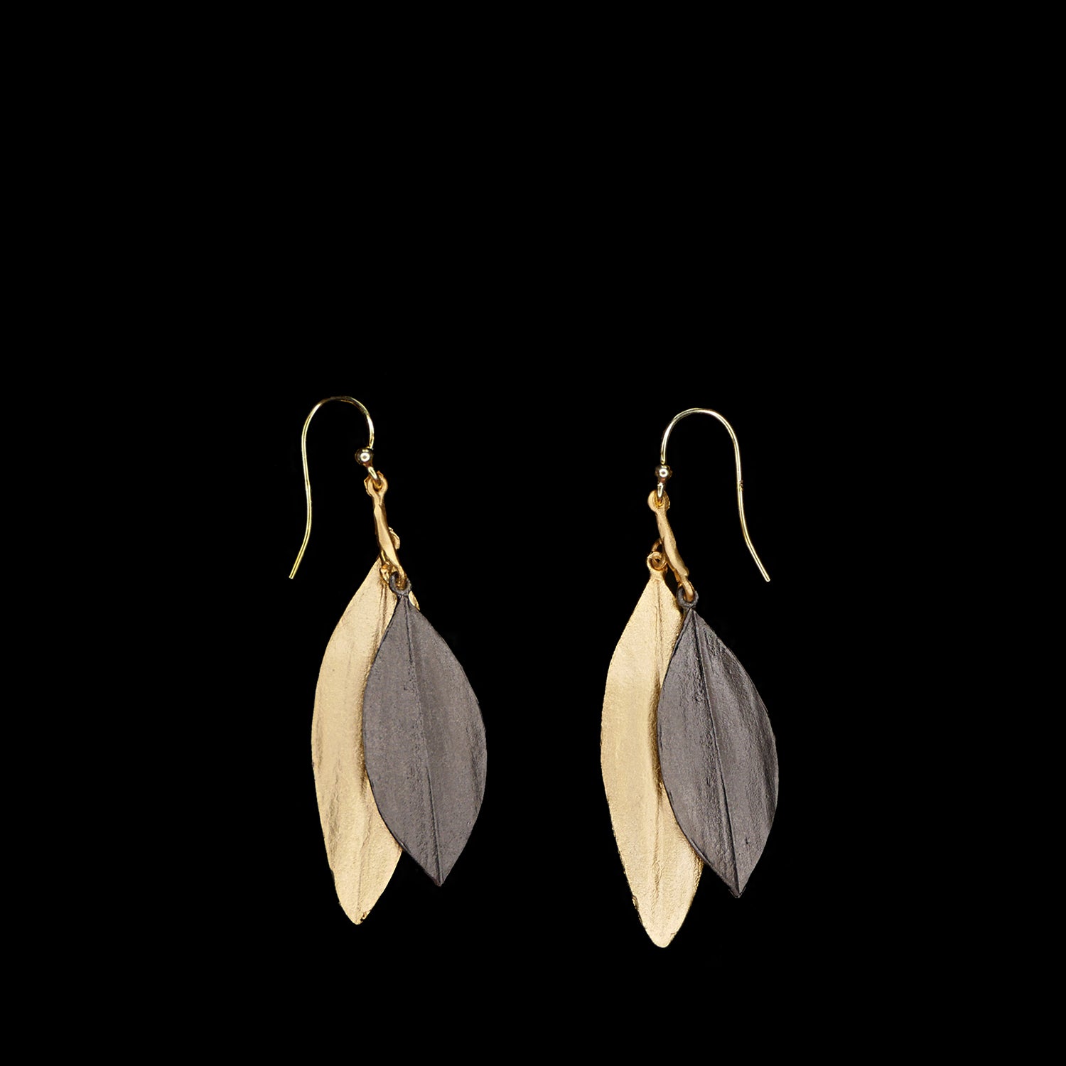 A pair of dangle earrings with overlapping gold and dark gray leaf-shaped metal charms against a black background.