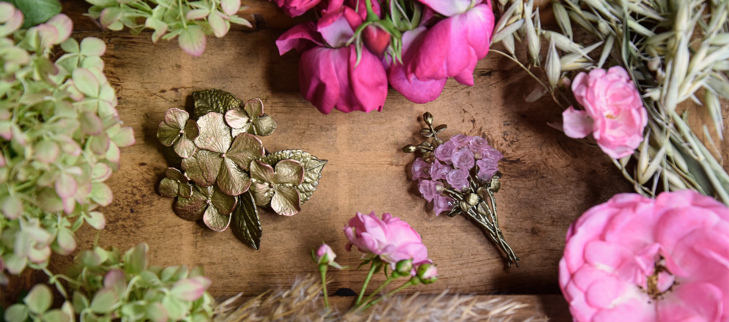 Two floral brooches, one gold and one with pink accents, are placed on a wooden surface surrounded by pink and green flowers.