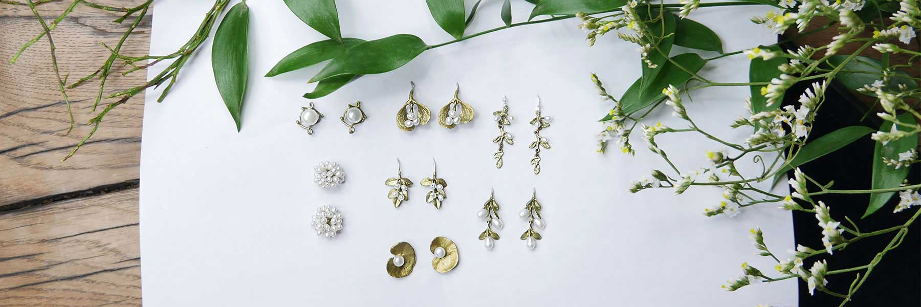A display of ten pairs of gold and pearl earrings arranged on white paper, surrounded by green leaves and small white flowers on a wooden surface.