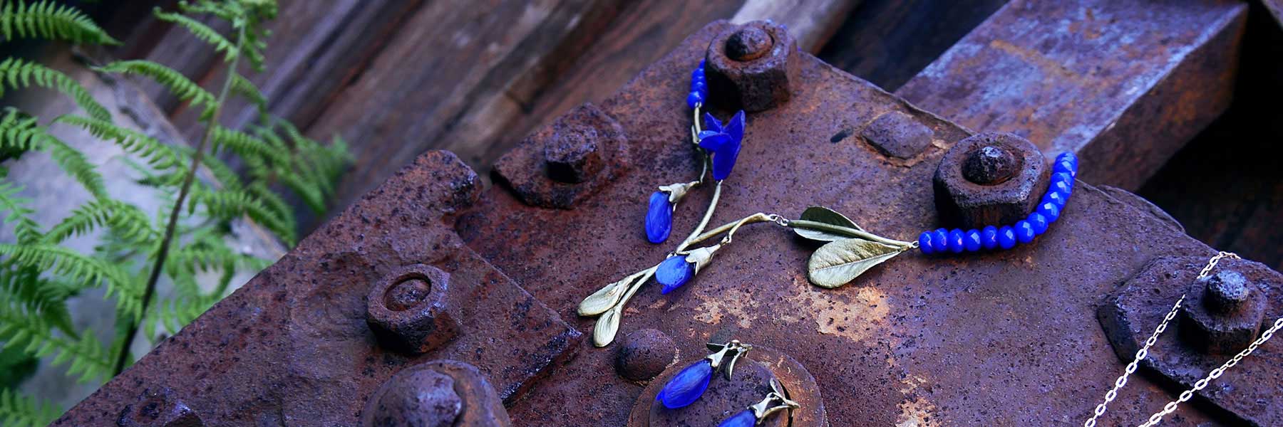 Blue and gold jewelry pieces are displayed on a rusty metal surface with a green fern visible in the background.