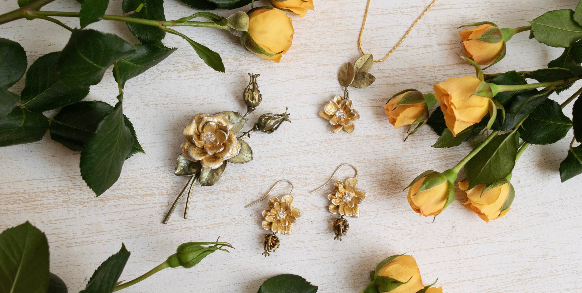 Gold floral jewelry including a brooch, necklace, and earrings arranged on a wooden surface with yellow rose flowers and green leaves.