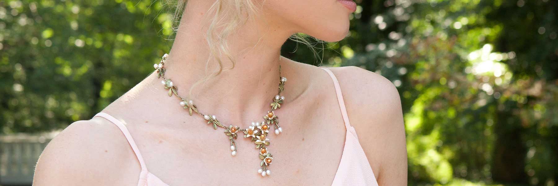 A woman wearing a pale pink top and a floral necklace with pearl and gold accents, outdoors with greenery in the background.
