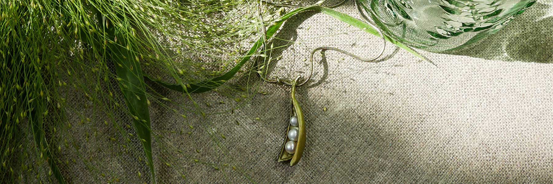 A gold pea pod necklace with three pearls rests on a textured beige fabric, surrounded by green foliage and a glass object.