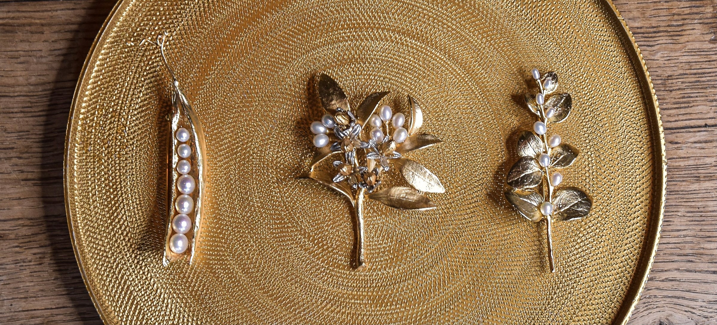 Three gold brooches with pearl details, shaped like a pea pod, a branch with leaves, and a sprig with round leaves, displayed on a textured gold plate.