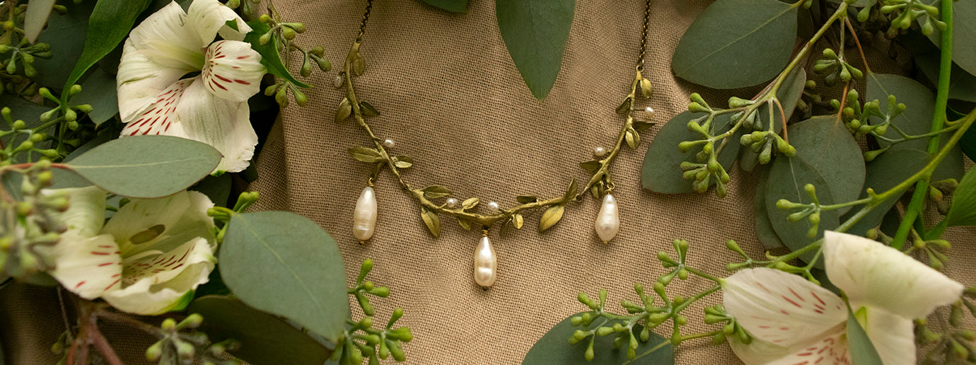A gold necklace with pearl pendants is displayed on a tan fabric, surrounded by green leaves and white flowers.