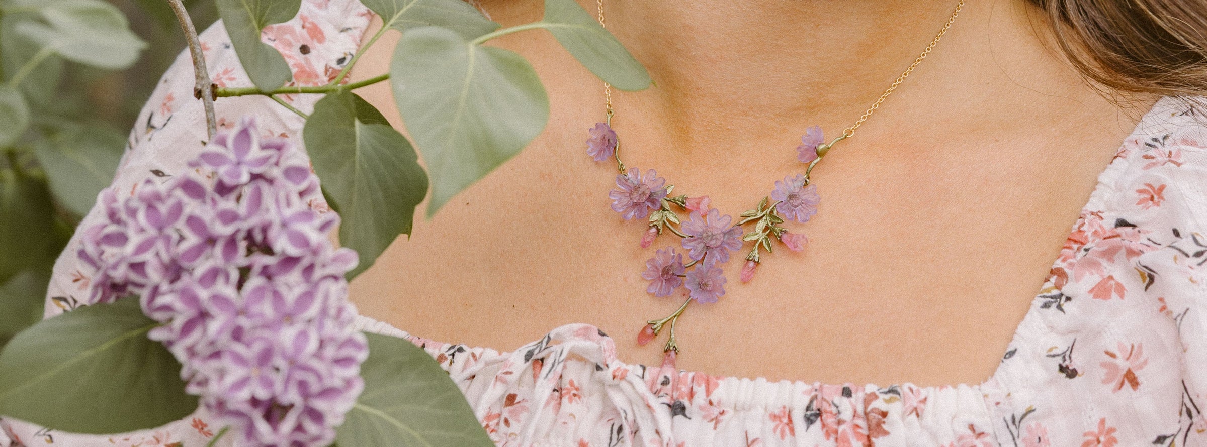 A person wearing a floral-patterned dress and a gold chain necklace with purple flower charms, standing near lilac flowers.