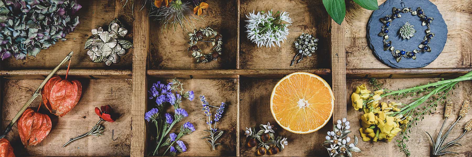 A flat lay of vintage jewelry and natural elements including dried flowers, fruit, and seed pods arranged in a wooden tray.