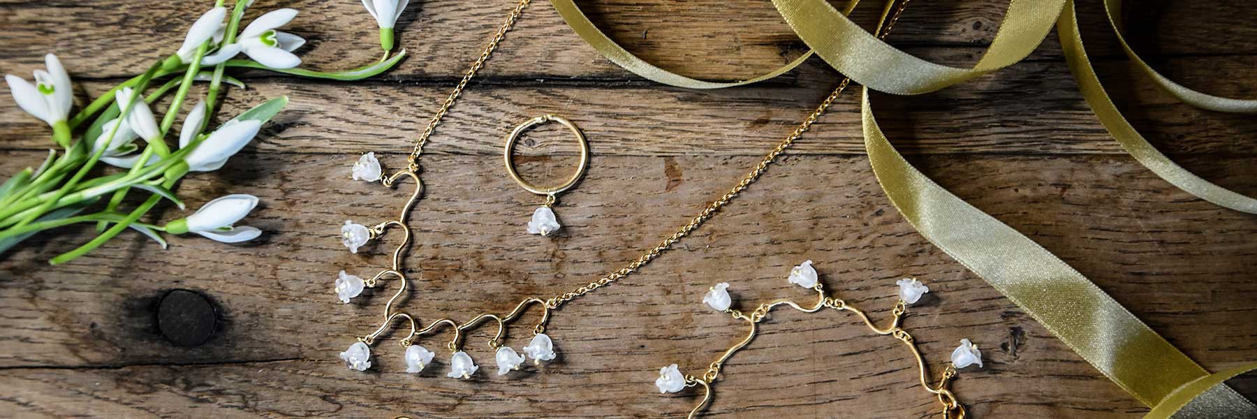 Gold jewelry set with white flower-shaped accents displayed on a wooden surface beside pale green ribbon and white flowers.