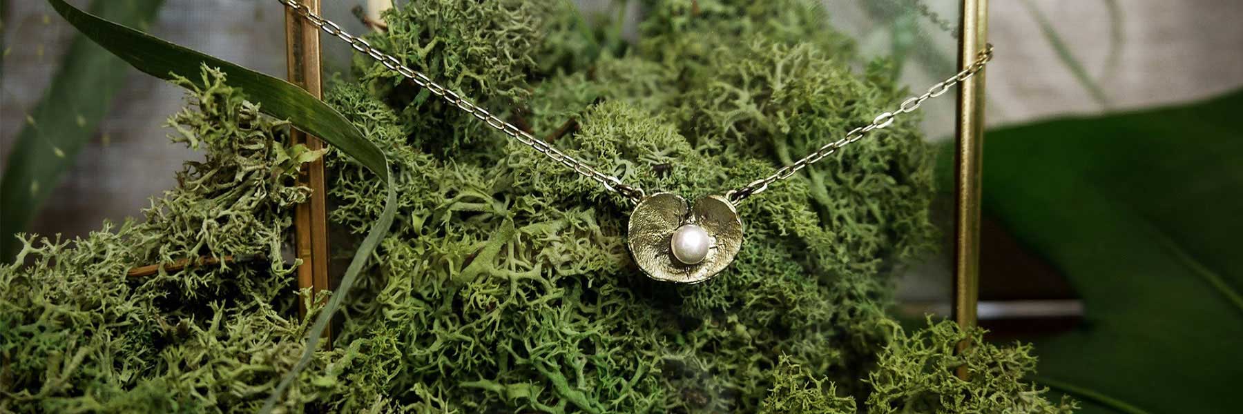 A silver necklace with a pendant shaped like a flower holding a pearl is displayed on green moss with leaves in the background.