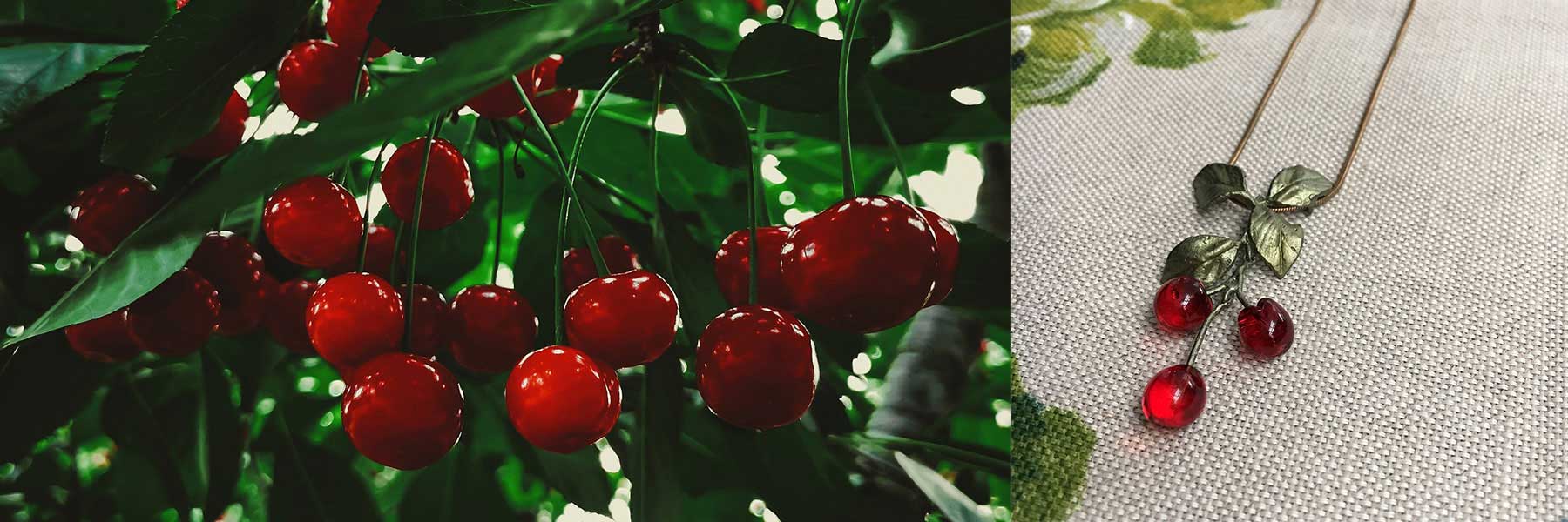 A cluster of red cherries hanging on a tree branch beside a necklace with cherry-shaped pendants on a textured fabric surface.