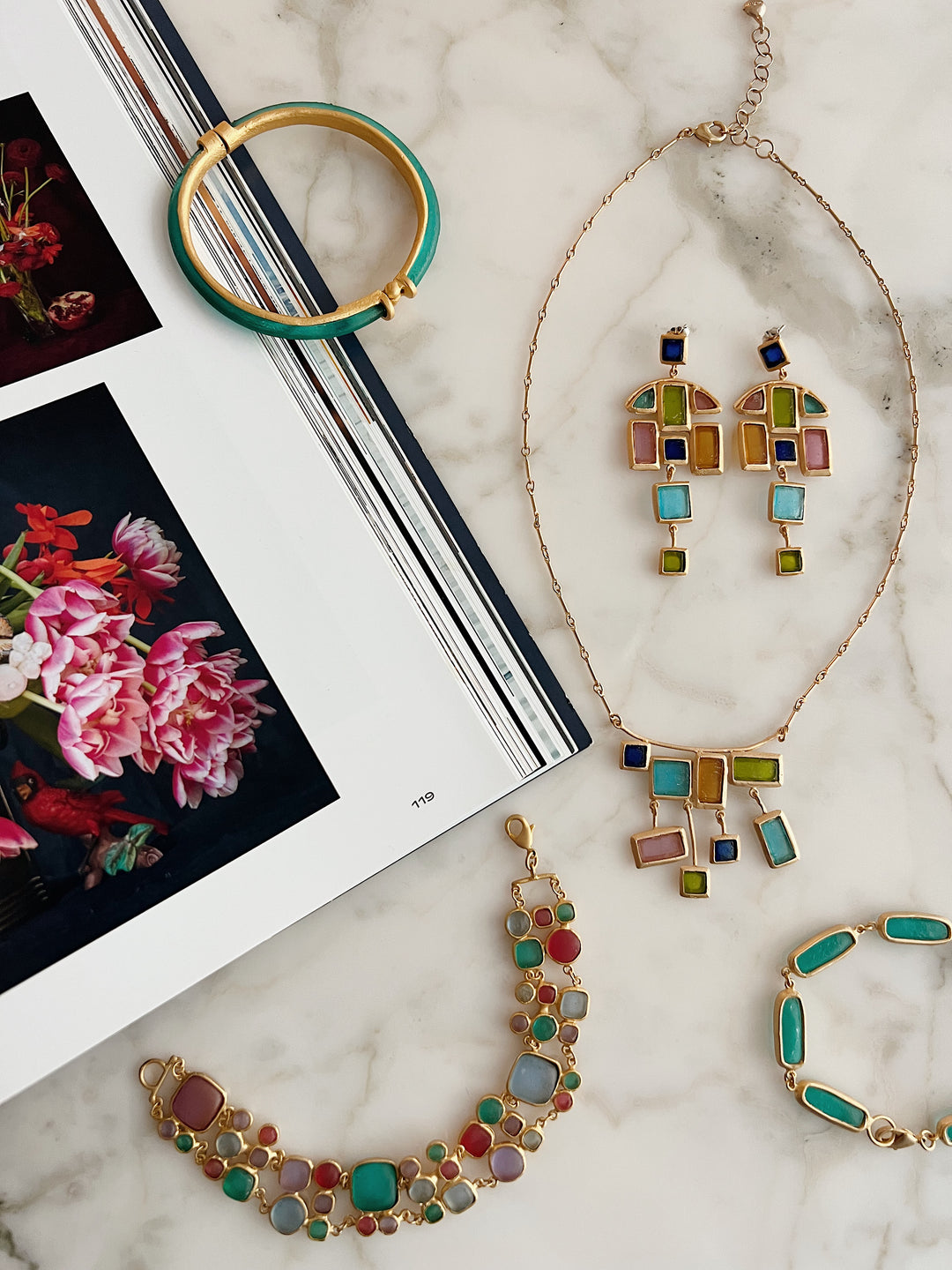A flat lay of colorful glass jewelry, including a necklace, bracelet, earrings, and two bangles, arranged on a marble surface next to an open book displaying floral photographs.