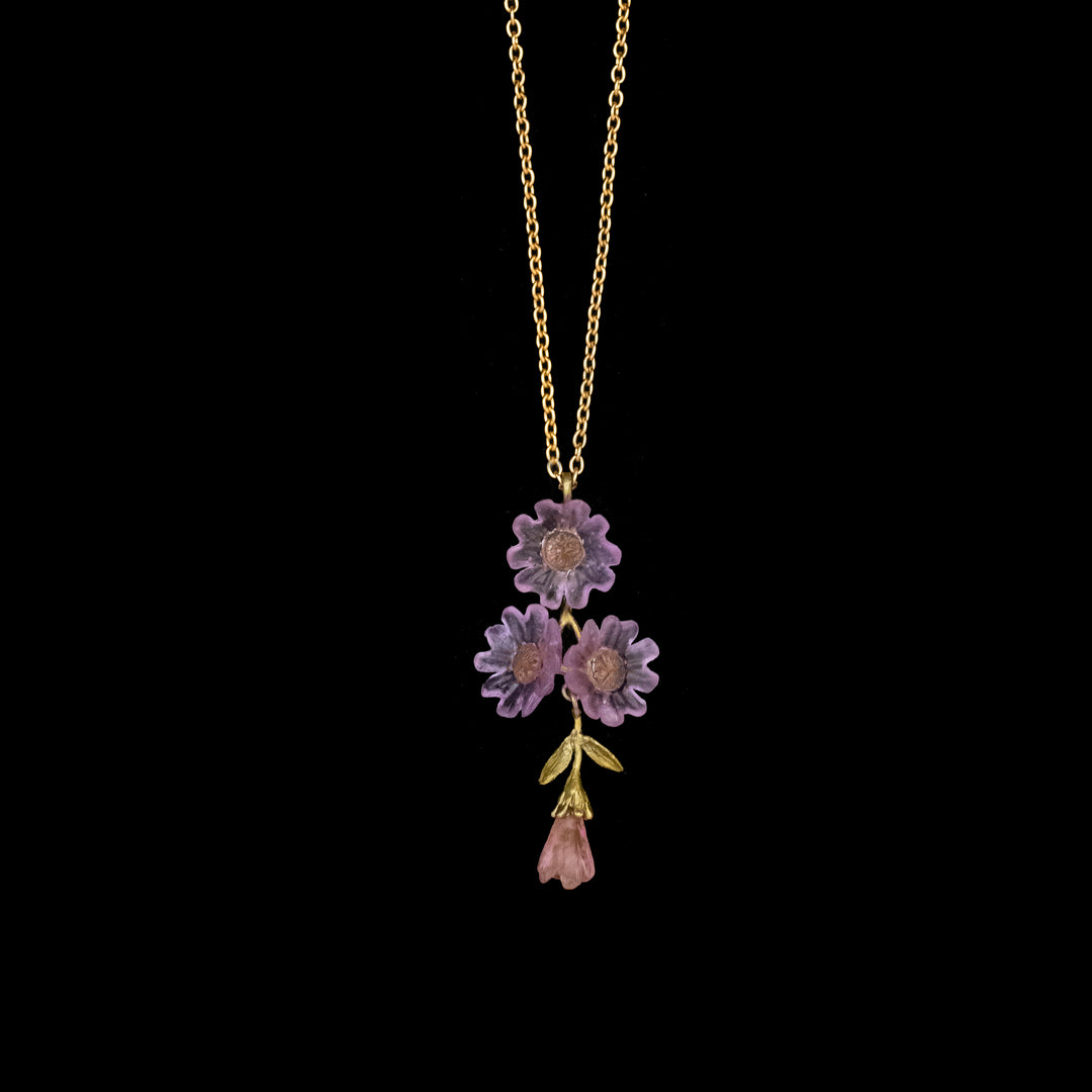 A gold necklace features a pendant with three purple flower shapes, green leaves, and a pink glass buds, displayed on a black background.