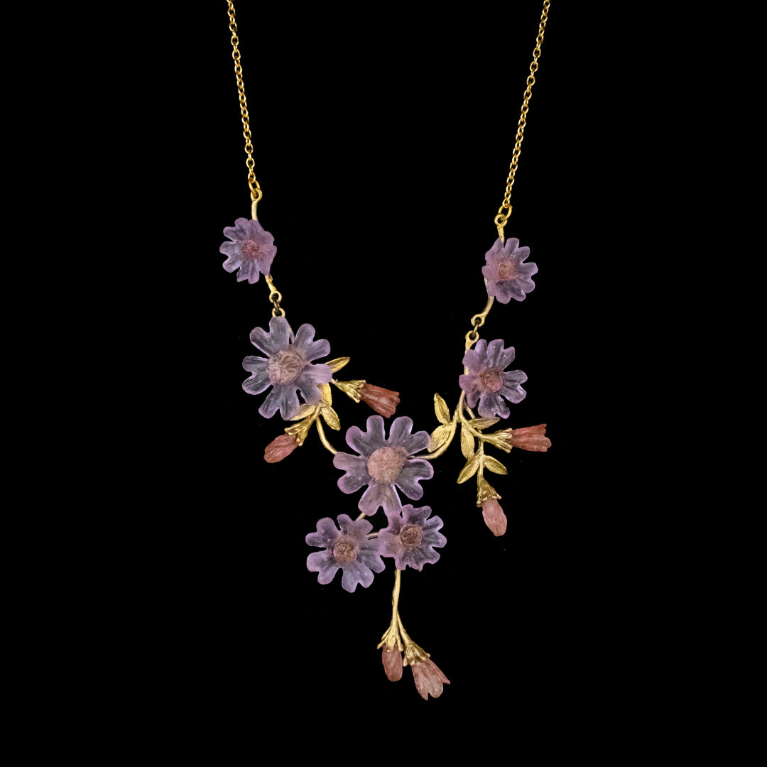 Gold necklace with purple and pink flower-shaped ornaments arranged along the chain and branch-like green accents, set against a black background.