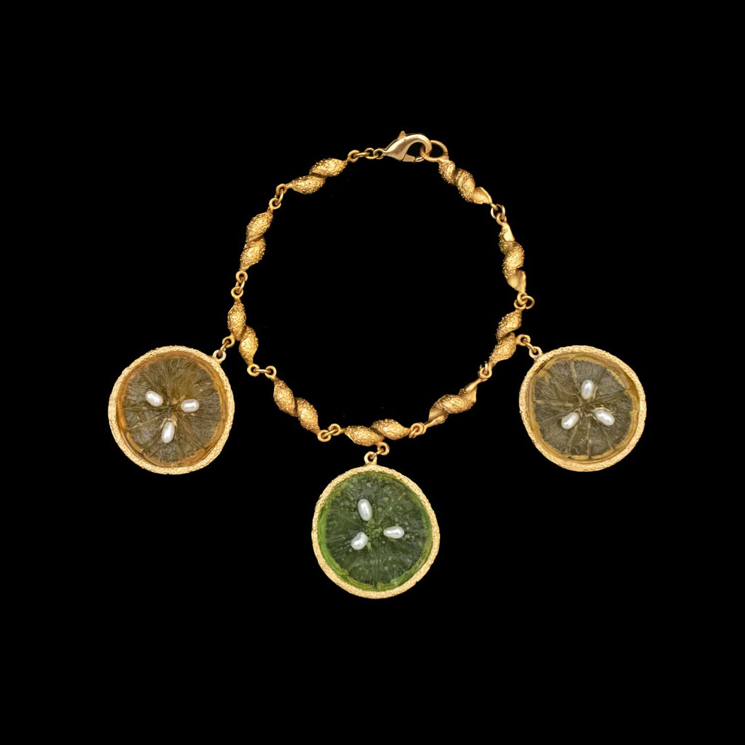 The Citrus Bracelet by Michael Michaud Jewelry features three 24kt gold plated circular charms resembling green, yellow and orange citrus fruit slices with visible seeds, all set against a black background.