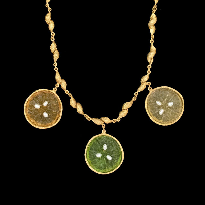 The Citrus Necklace by Michael Michaud Jewelry features three handcrafted, gold-plated circular pendants in yellow, green, and orange with white seed pearls, evoking citrus fruit slices on a black background.