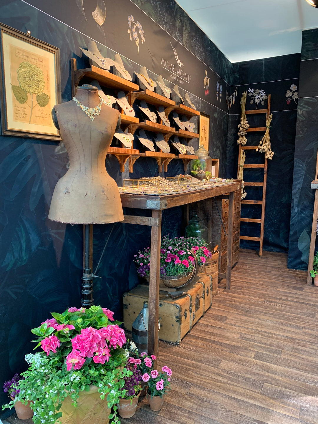 A display area with a wooden table holding jewelry, a vintage dress form, shelves with necklaces, framed art on the wall, a wooden ladder, and potted flowers on the floor and trunks, all set on a wood floor.