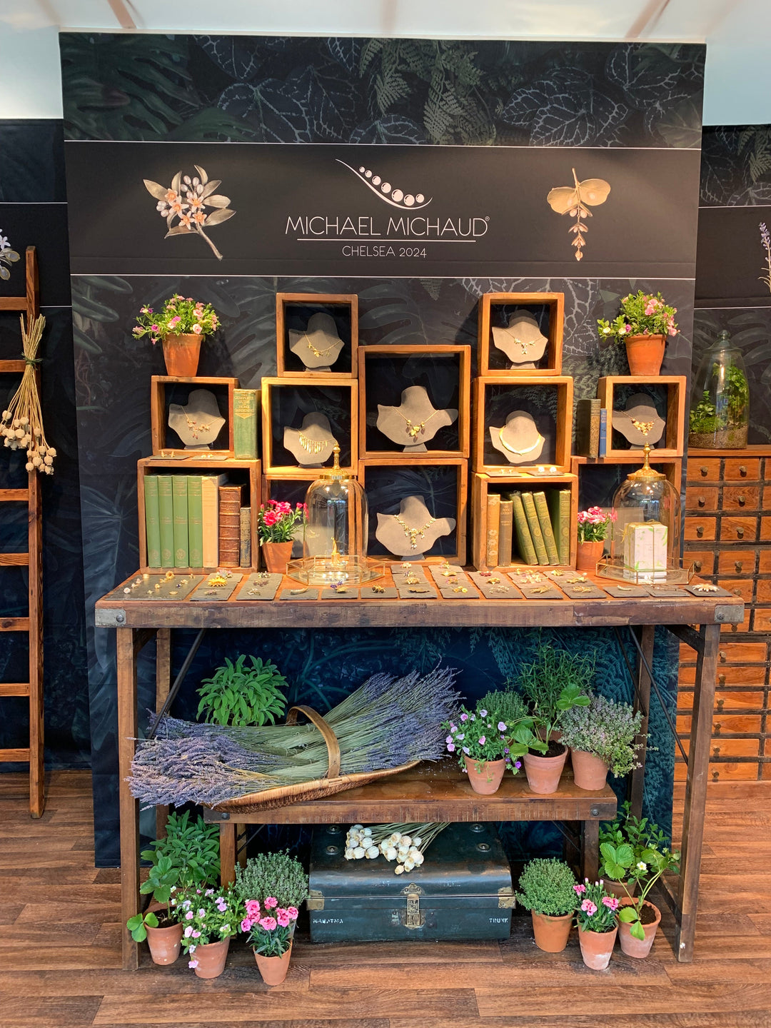 A wooden table with potted plants and lavender bundles is in front of a display of jewelry on stands, set in wooden boxes. The backdrop reads “Michael Michaud Chelsea 2024,” with botanical decorations.
