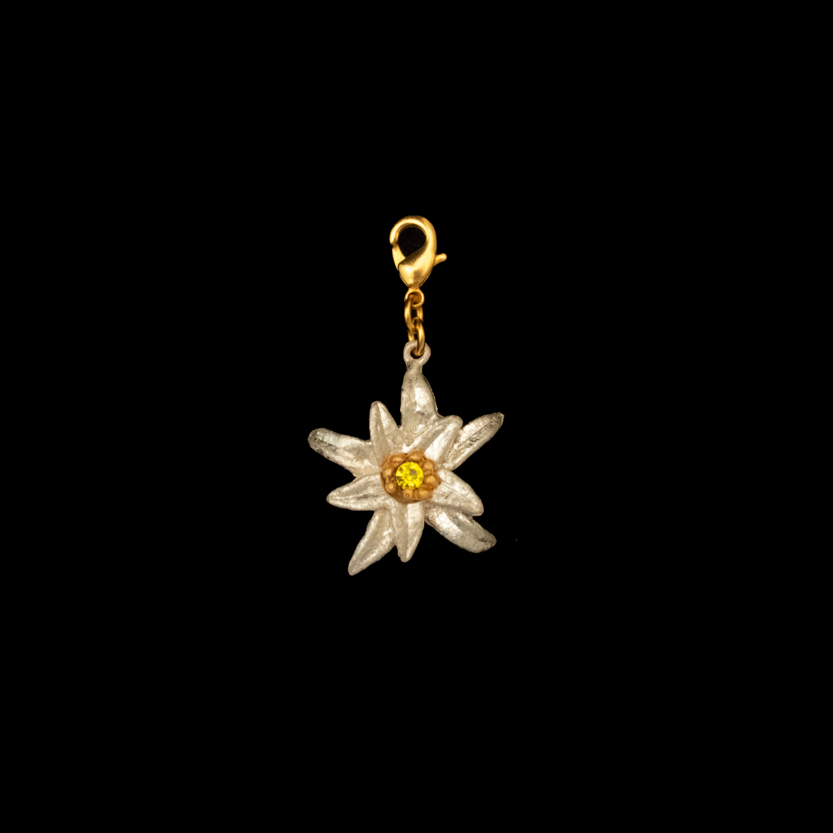 The Edelweiss 60th Anniversary Commemorative Charm, inspired by The Sound of Music, features a white bloom with a yellow center on a gold lobster clasp and is shown against a black background.