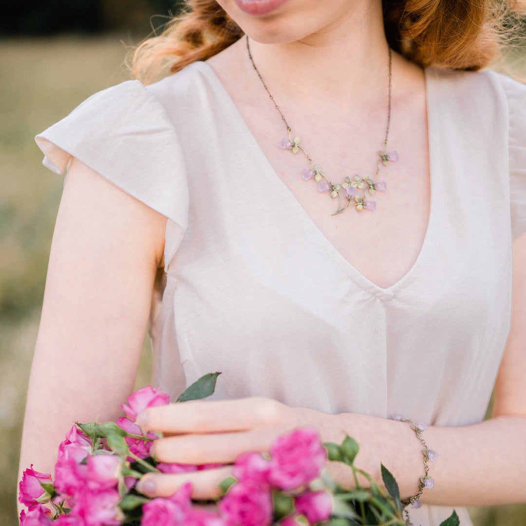 Woman in a light-colored dress holds a bouquet of pink flowers and wears a rose necklace and bracelet, standing outdoors—perfect for Valentine's Day.