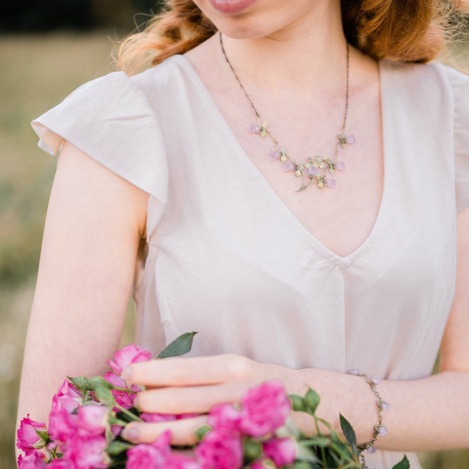 Woman in a light-colored dress holds a bouquet of pink flowers and wears a rose necklace and bracelet, standing outdoors—perfect for Valentine's Day.