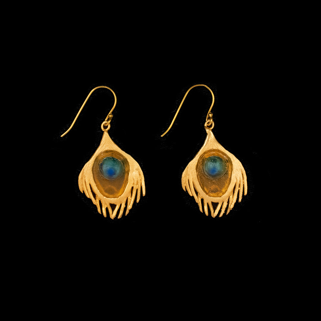 A pair of gold-colored bronze earrings with a peacock feather design, featuring a blue accent, displayed against a black background.