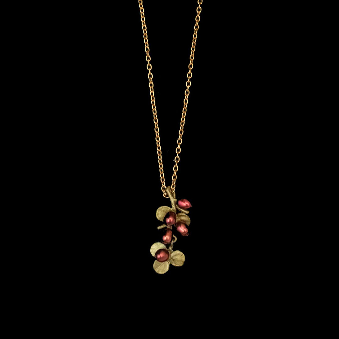 The Firethorn Pendant is a gold chain necklace featuring green leaves and red freshwater pearls, shown against a solid black background—a striking piece of botanical jewelry.