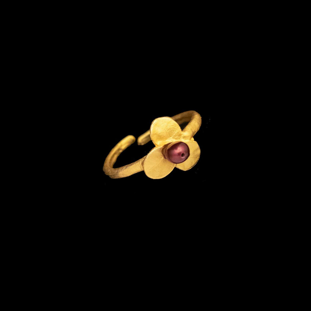The Firethorn Adjustable Ring is gold-plated with an open band and a leaf-shaped top featuring three gold petals and a round red freshwater pearl at the center, shown against a black background.