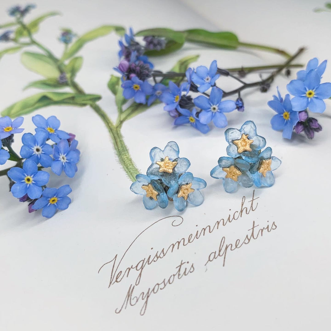 Blue and gold Forget Me Not Earrings are displayed on a page with botanical drawings and text reading Vergissmeinnicht Myosotis alpestris, surrounded by real blue forget-me-not flowers—a thoughtful piece from our Mother’s Day collection.