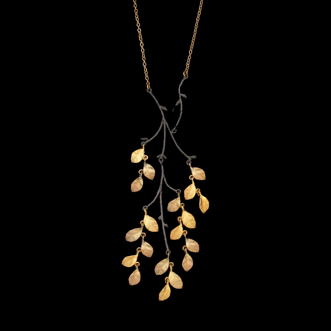 The Golden Autumn Necklace features a gold chain with a pendant of tree-branch shapes, decorated with gold and antique bronze birch leaves on black metal stems, all set against a black background.