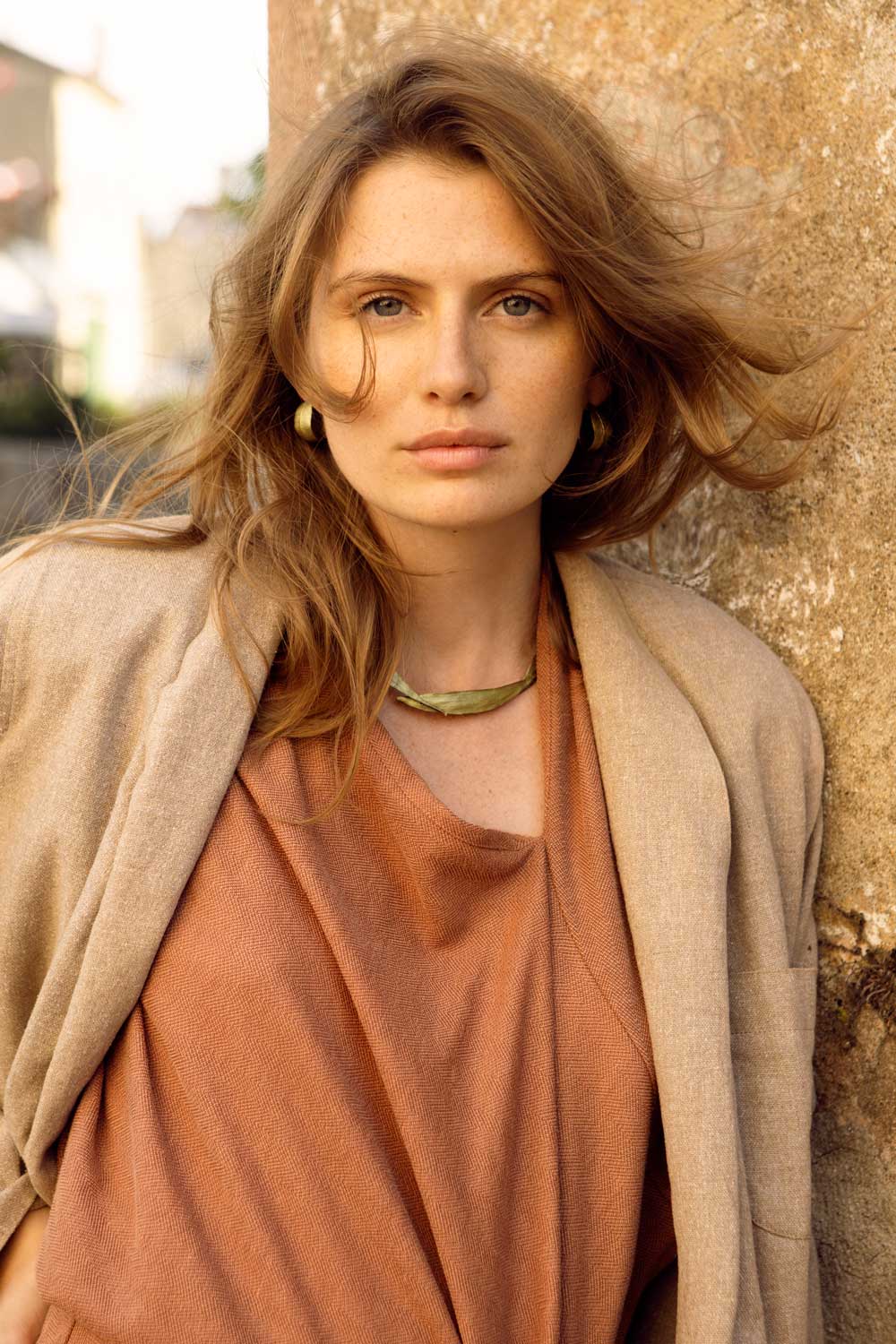 A woman with light brown hair wears a tan blazer over a rust-colored top, standing against a weathered stone wall outdoors. She has a neutral expression and is looking directly at the camera.