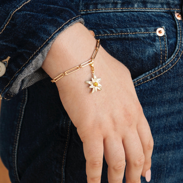 A person wearing a gold chain bracelet with the Edelweiss 60th Anniversary Commemorative Charm, dressed in blue denim jeans and jacket, rests their hand on their hip.