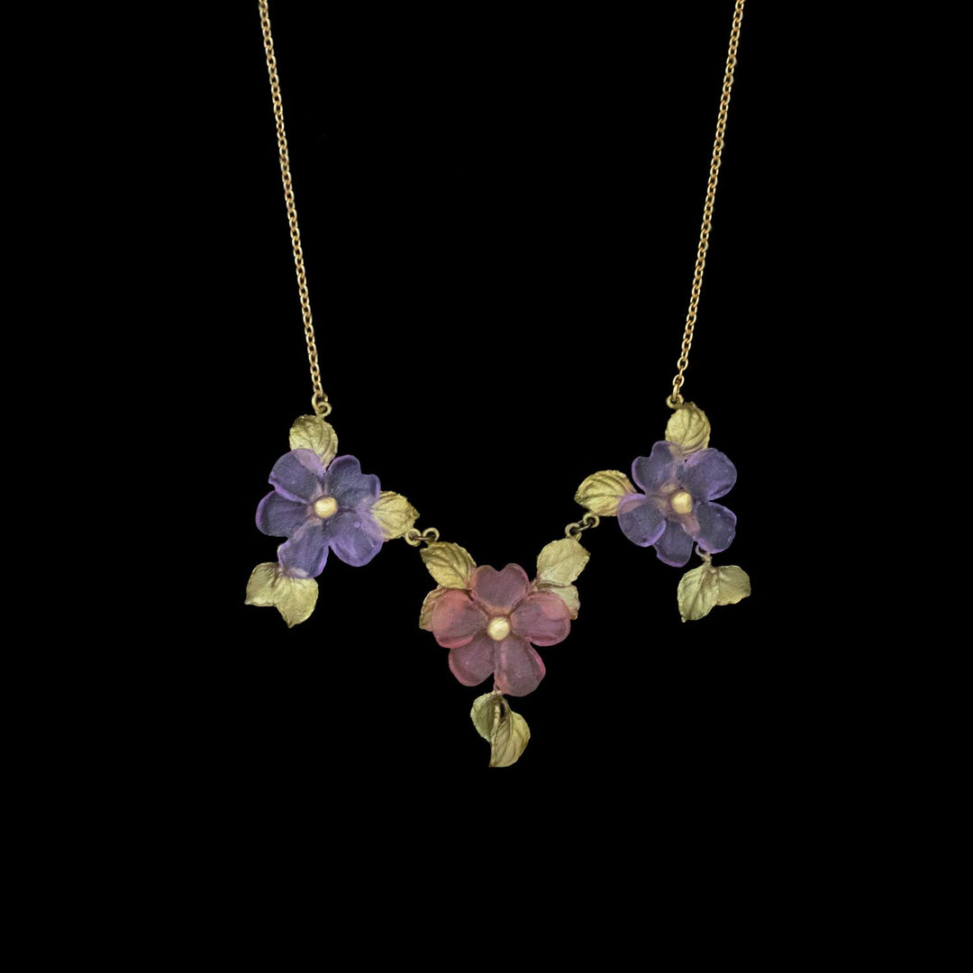Handcrafted floral necklace featuring three flower pendants—two purple and one pink—accented by gold leaves, this Impatiens jewelry piece captures the beauty of nature-inspired accessories against a black background.