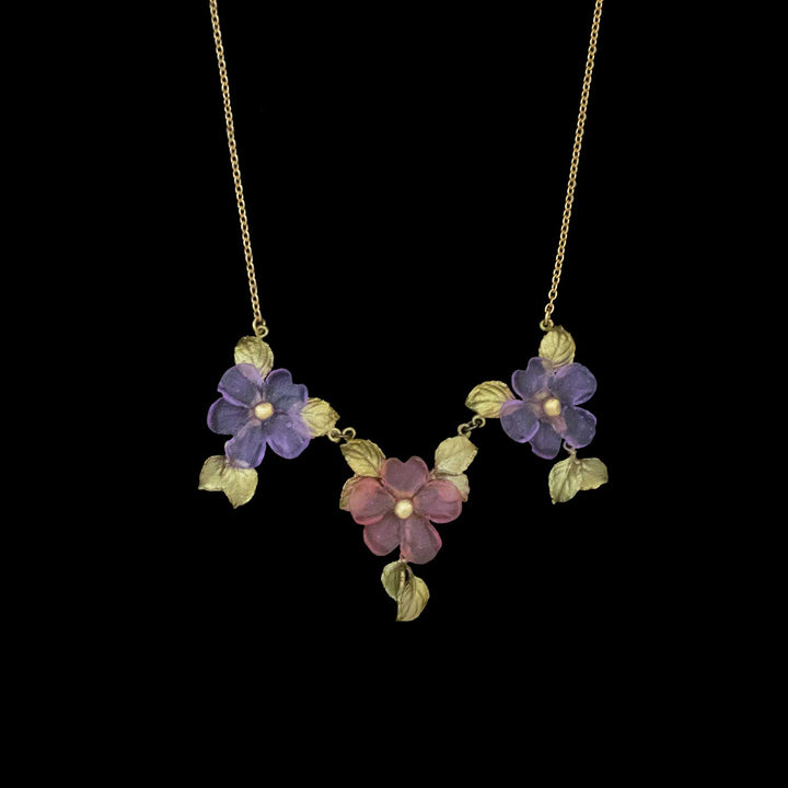 Handcrafted floral necklace featuring three flower pendants—two purple and one pink—accented by gold leaves, this Impatiens jewelry piece captures the beauty of nature-inspired accessories against a black background.