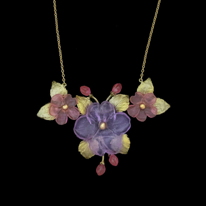 Handcrafted floral necklace featuring a central purple flower with golden center, flanked by two pink flowers and green leaves, accented with small pink buds, on a black background—an elegant piece for nature-inspired accessories lovers.