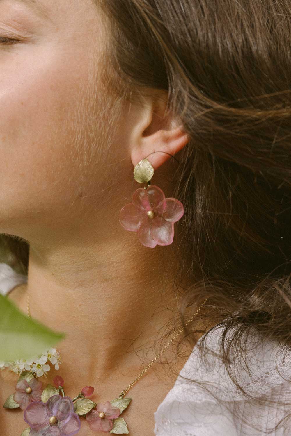 A close-up of a woman wearing handcrafted flower earrings and a matching floral necklace. She is dressed in a white top, her brown hair loose, showcasing elegant, nature-inspired accessories.