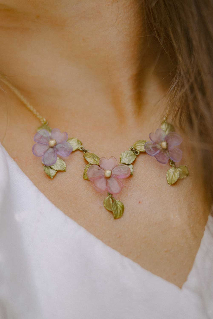 A close-up of a handcrafted floral necklace featuring three pastel flower pendants with green leaves and pearl centers, worn with a white top—a beautiful example of nature-inspired accessories.