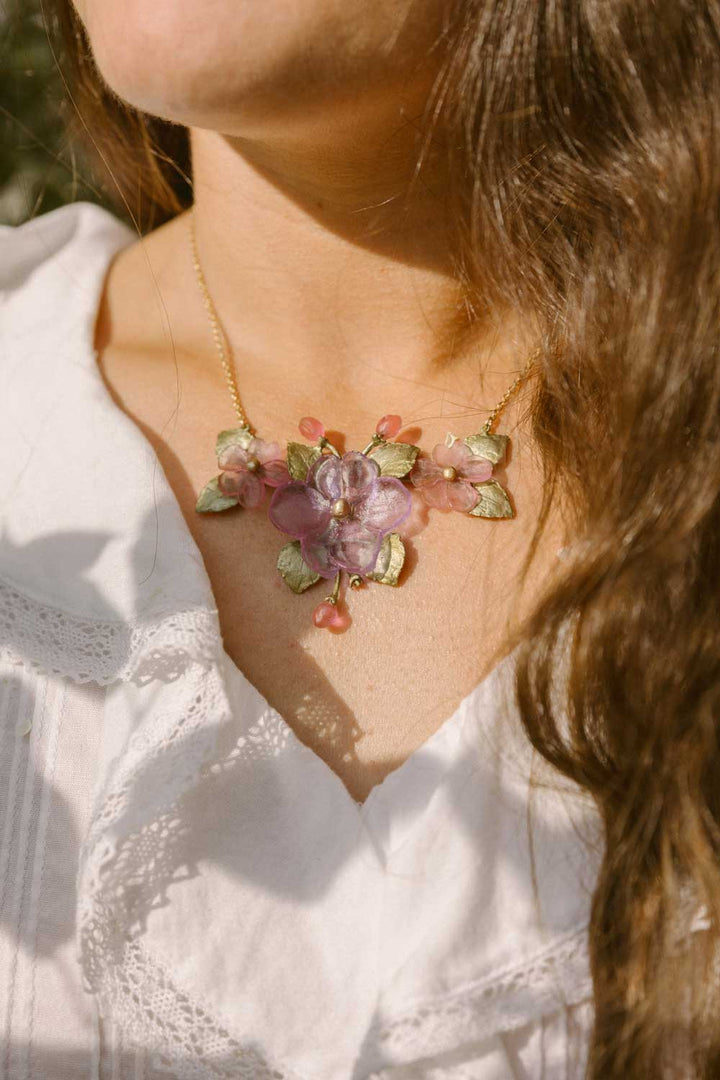 A person wearing a white shirt and a gold, handcrafted floral necklace adorned with pink and green decorations, showcasing nature-inspired accessories.