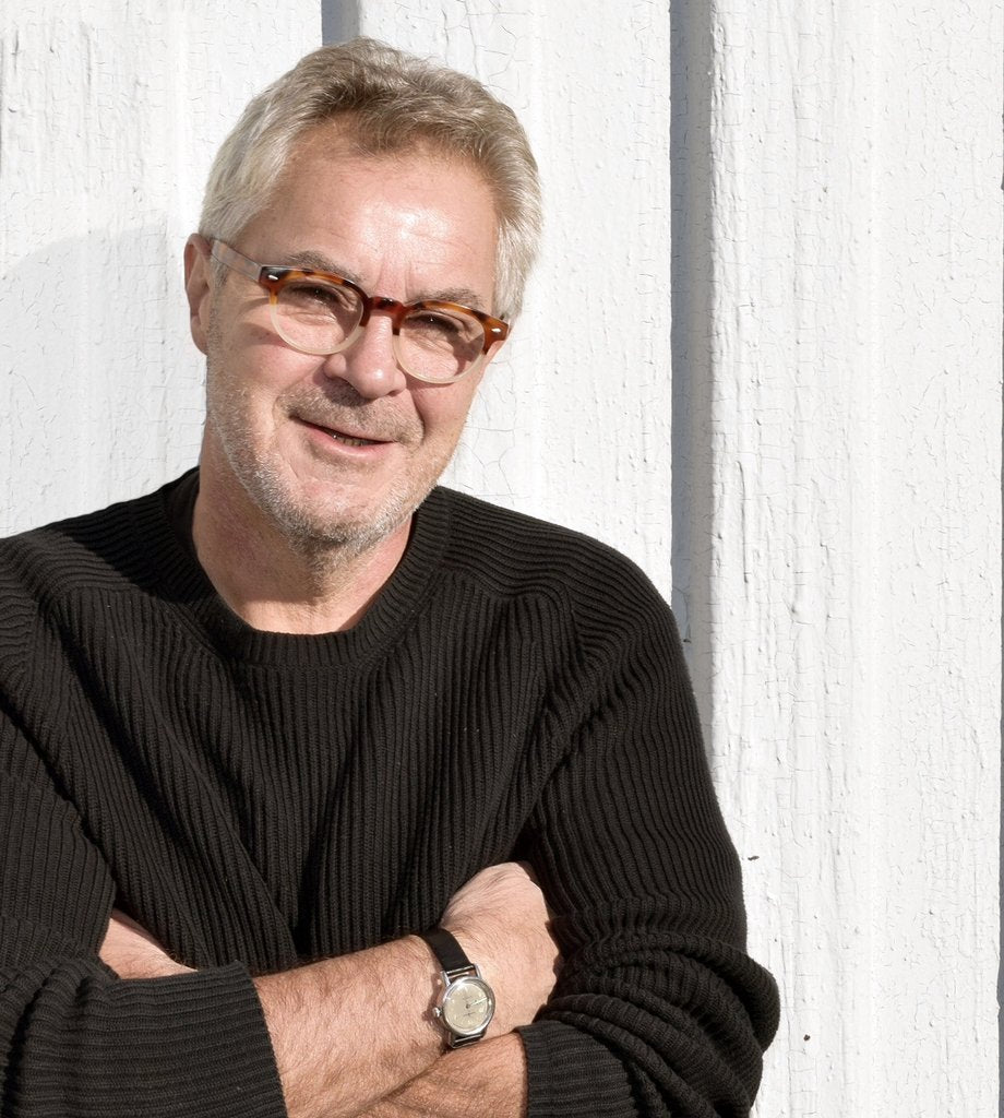 A middle-aged man with gray hair and glasses stands in front of a white wooden wall, wearing a black sweater and a wristwatch, with his arms crossed and a slight smile on his face.