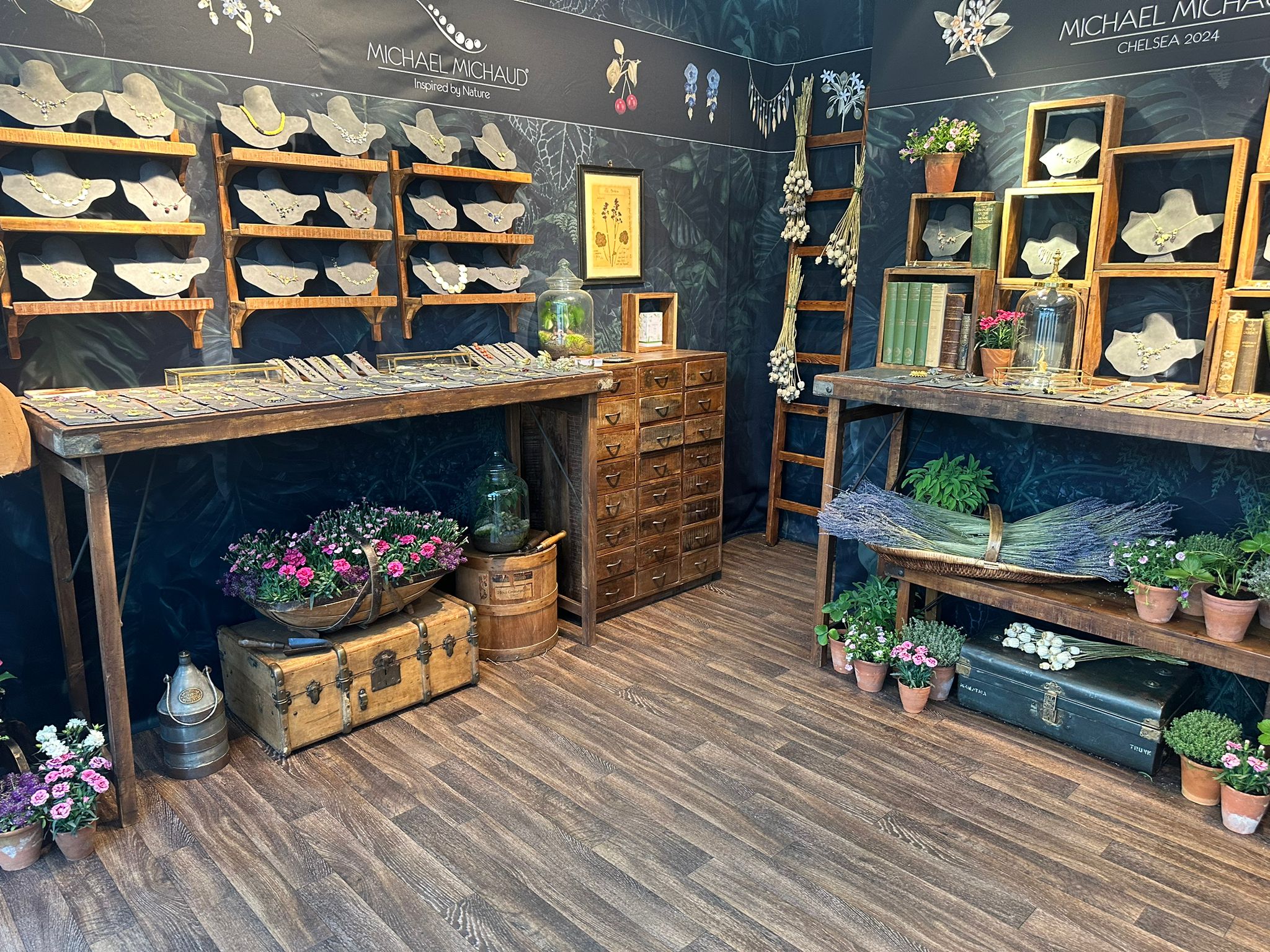 A boutique shop interior with wooden shelves displaying jewelry, tables with jewelry trays, potted plants, a ladder, vintage suitcases, a chest of drawers, and dried lavender bundles on wooden flooring.