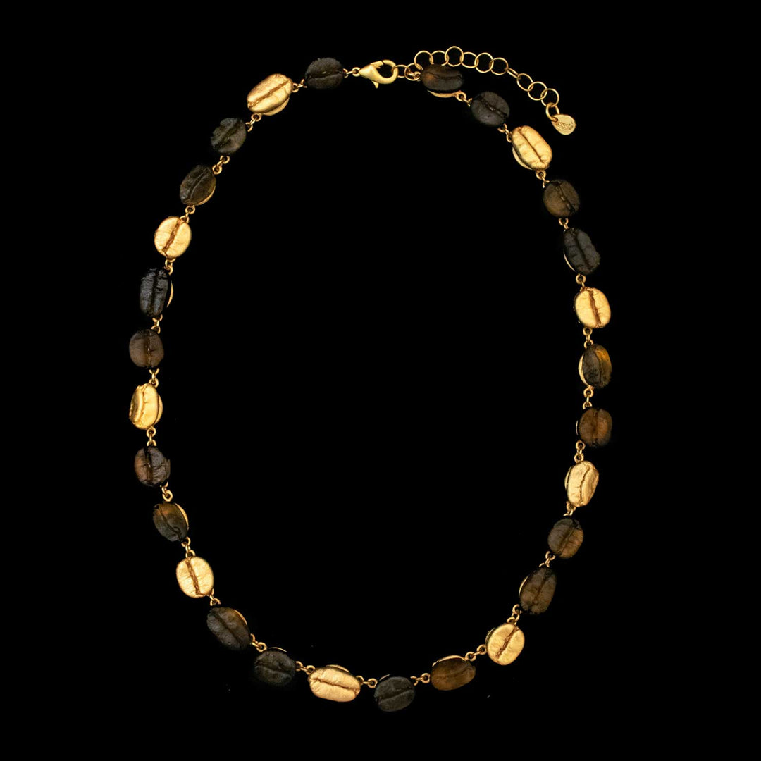 The Roasted Coffee Bean Necklace is gold-plated and features alternating gold and dark brown oval coffee beans, linked by small gold connectors, and showcased on a black background.