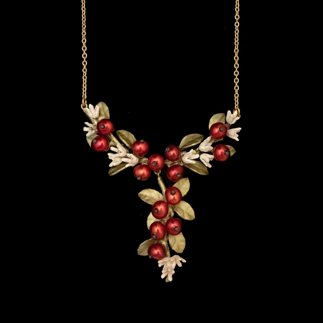 The Unroasted Coffee Bean Necklace is a gold chain piece with a pendant of green leaves  and white flower accents, red berry-like pearls, and hand-patinated bronze in a floral design, shown on a black background.