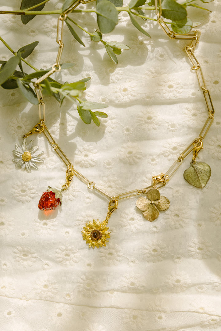 A gold jewelry chain necklace with charms shaped like a daisy, strawberry, sunflower, clover, and leaf, displayed on embroidered white fabric with green leaves above; features an extender for adjustable length.