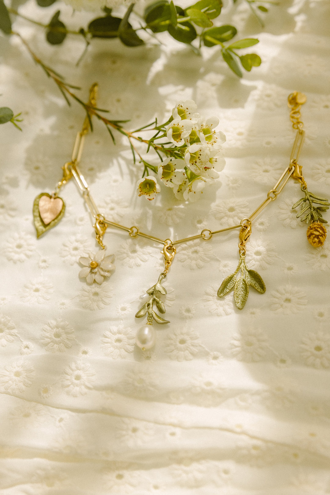 A gold charm bracelet with flower, leaf, and fruit charms is displayed on a white embroidered fabric, surrounded by small white flowers and green leaves, featuring an extender for the perfect jewelry fit.