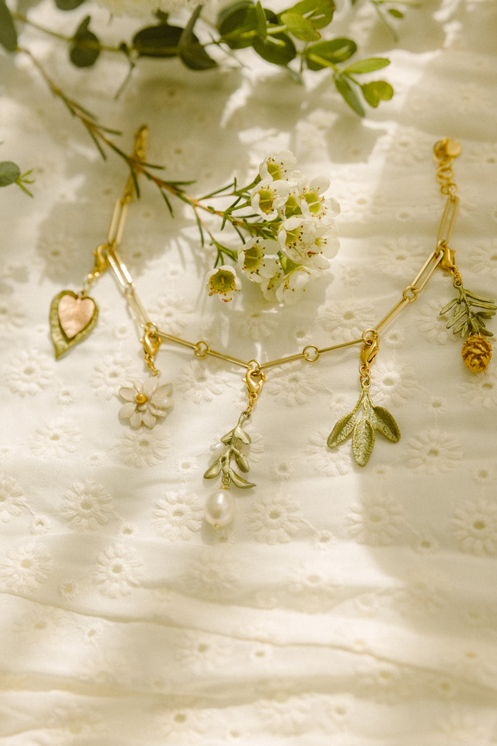 A gold charm bracelet with flower, leaf, and fruit charms is displayed on a white embroidered fabric, surrounded by small white flowers and green leaves, featuring an extender for the perfect jewelry fit.