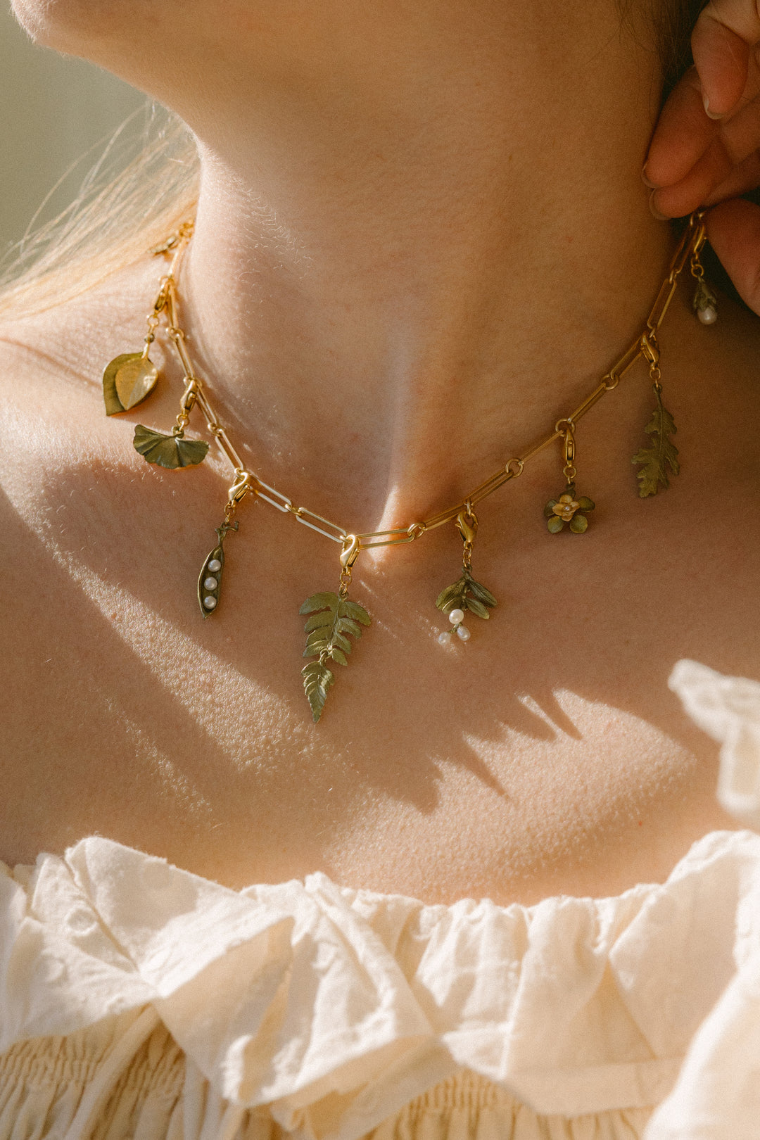 A close-up of a golden necklace with leaf and small charm pendants, worn by a person in a cream off-shoulder top. This jewelry piece features an extender for adjustable length.